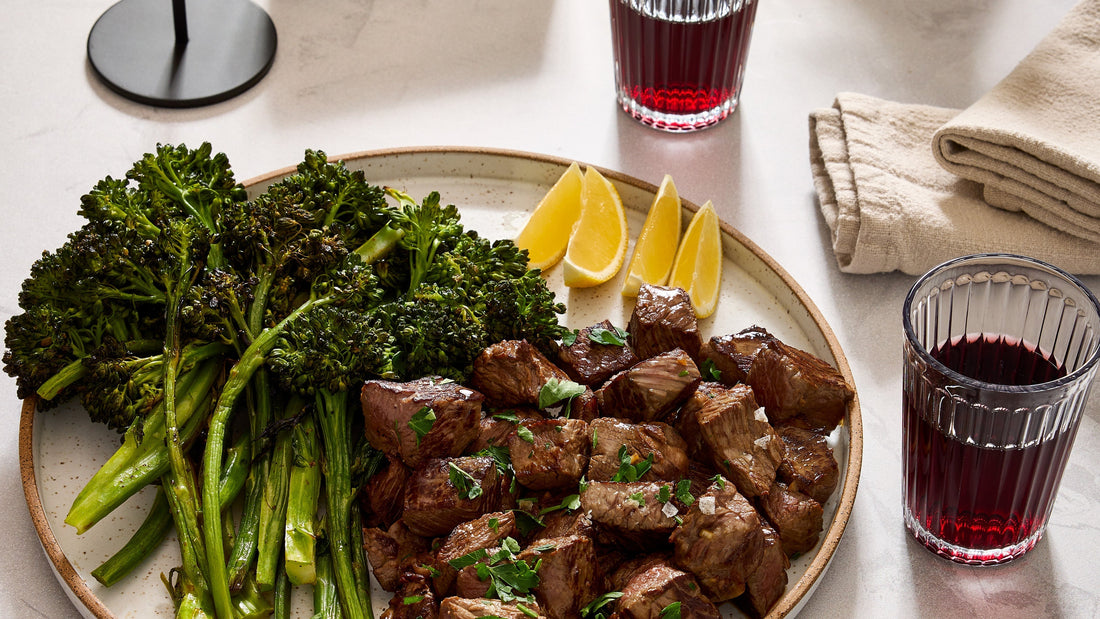 A dinner plate with Garlic-Ghee Steak Bites & Charred Broccolini next to a glass of red wine.