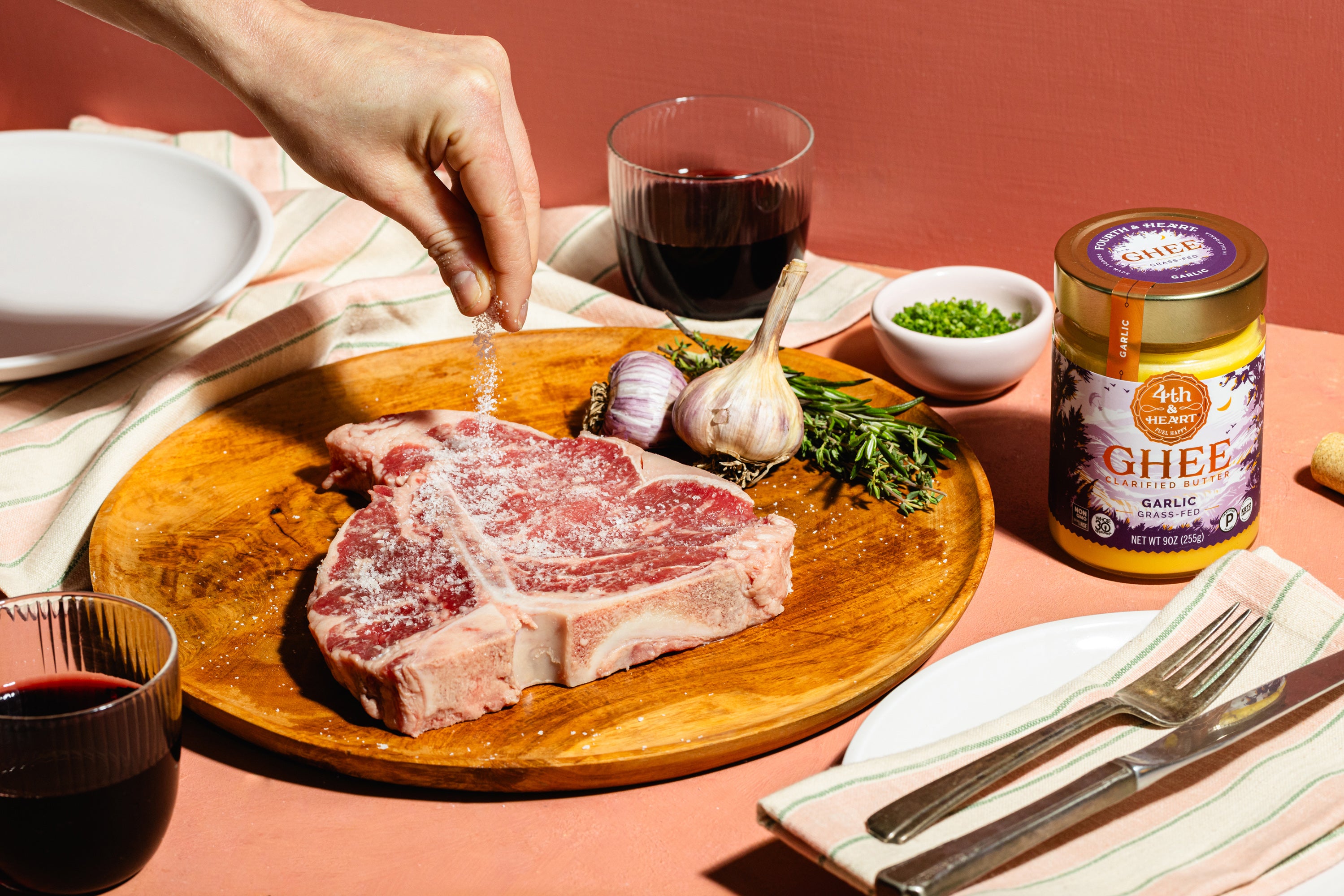 Person seasoning raw meat on a wooden board with a jar of 4th & Heart Garlic ghee and glass of red wine in the background.