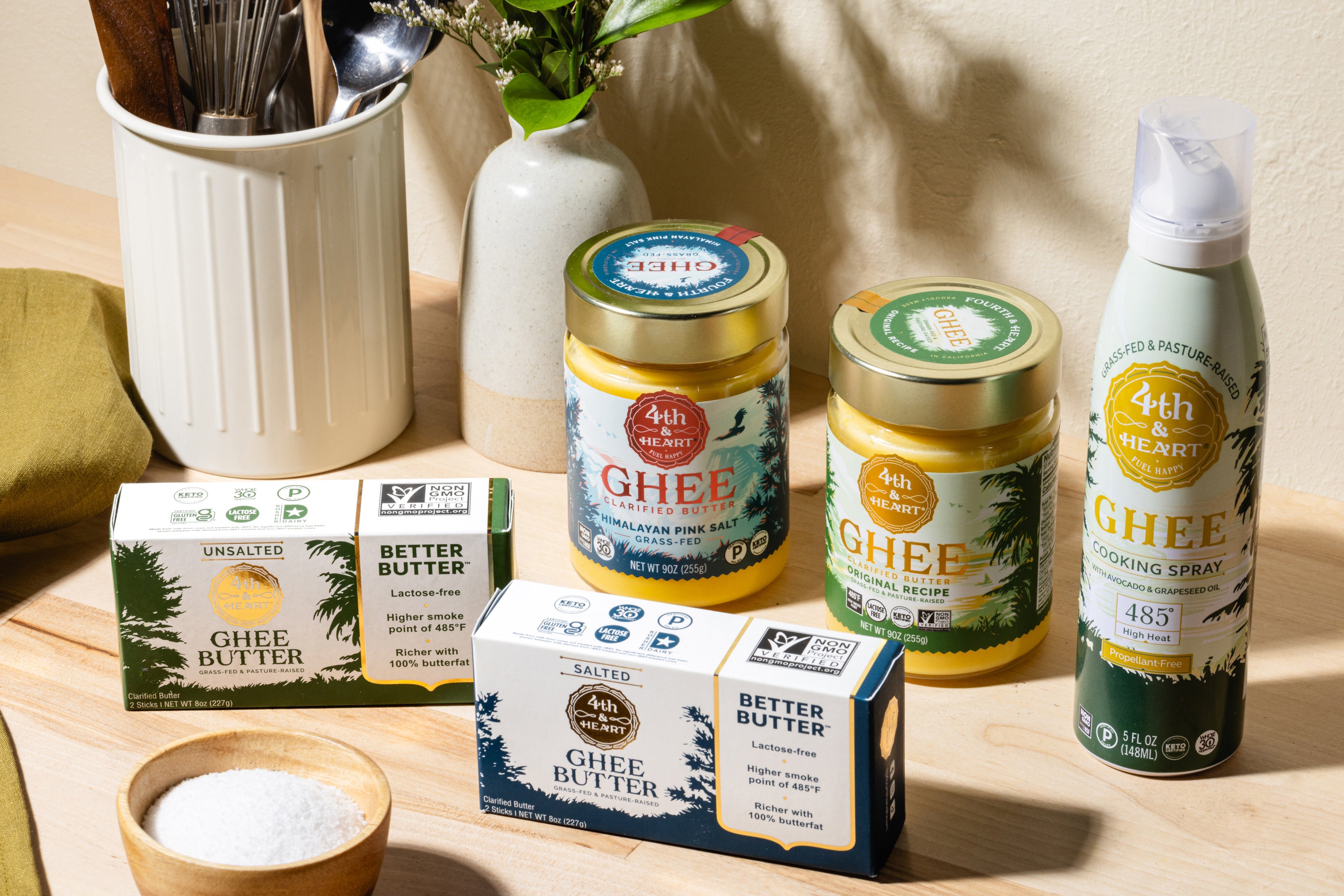 Assorted 4th & Heart Ghee products including clarified butter jars, butter stick boxes, and a cooking spray bottle on a wooden surface.